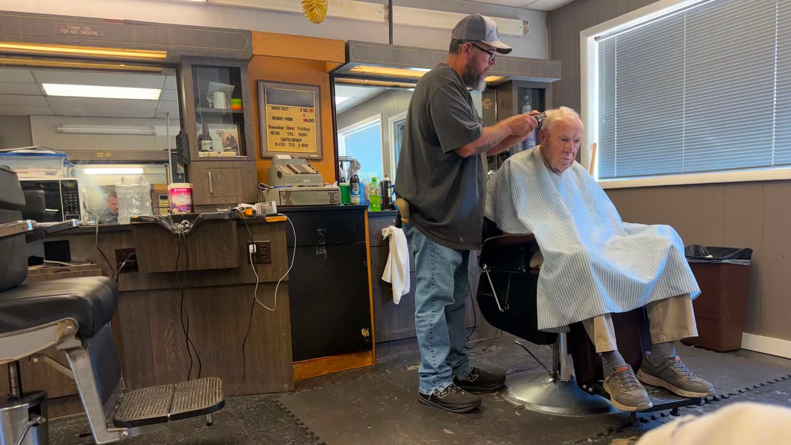 Chuck Haynie cutting a customer's hair inside Village Barber & Style Shop, the price board visible on the wall behind him.